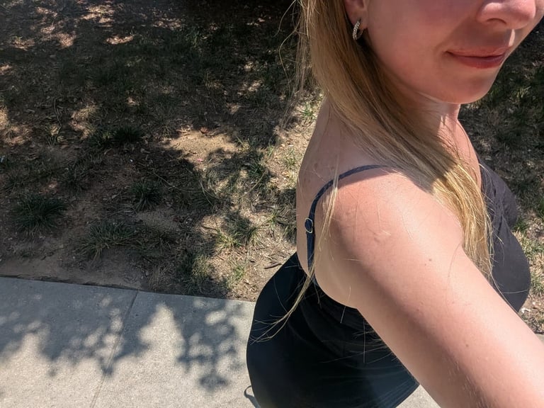 Woman with long blonde hair wearing a black sleeveless top, posing outdoors with tree shadows visible on ground behind her