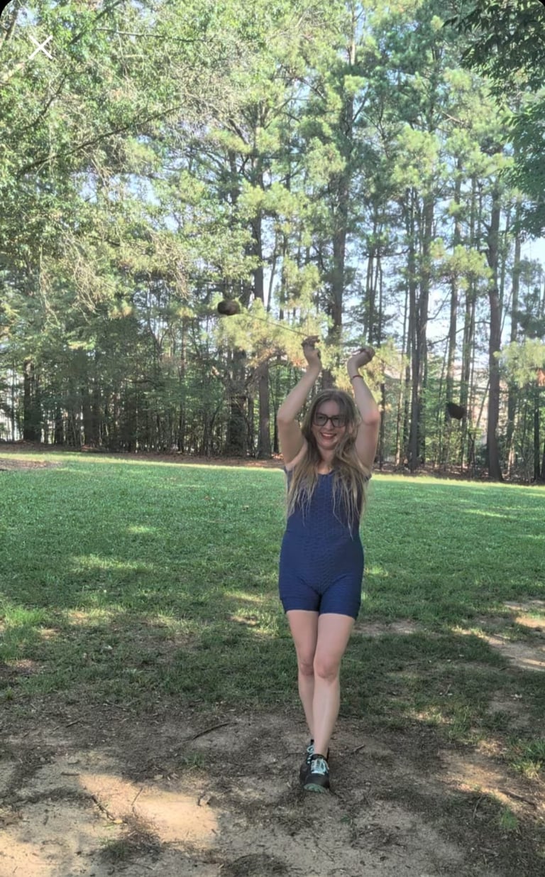 Woman in dark blue athletic romper with glasses stretching with arms raised on a grassy outdoor area surrounded by tall trees