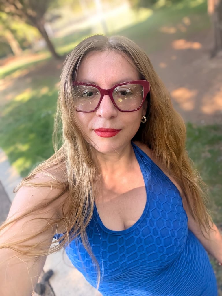 Woman with long reddish-brown hair wearing purple-framed glasses and a blue tank top, smiling at the camera