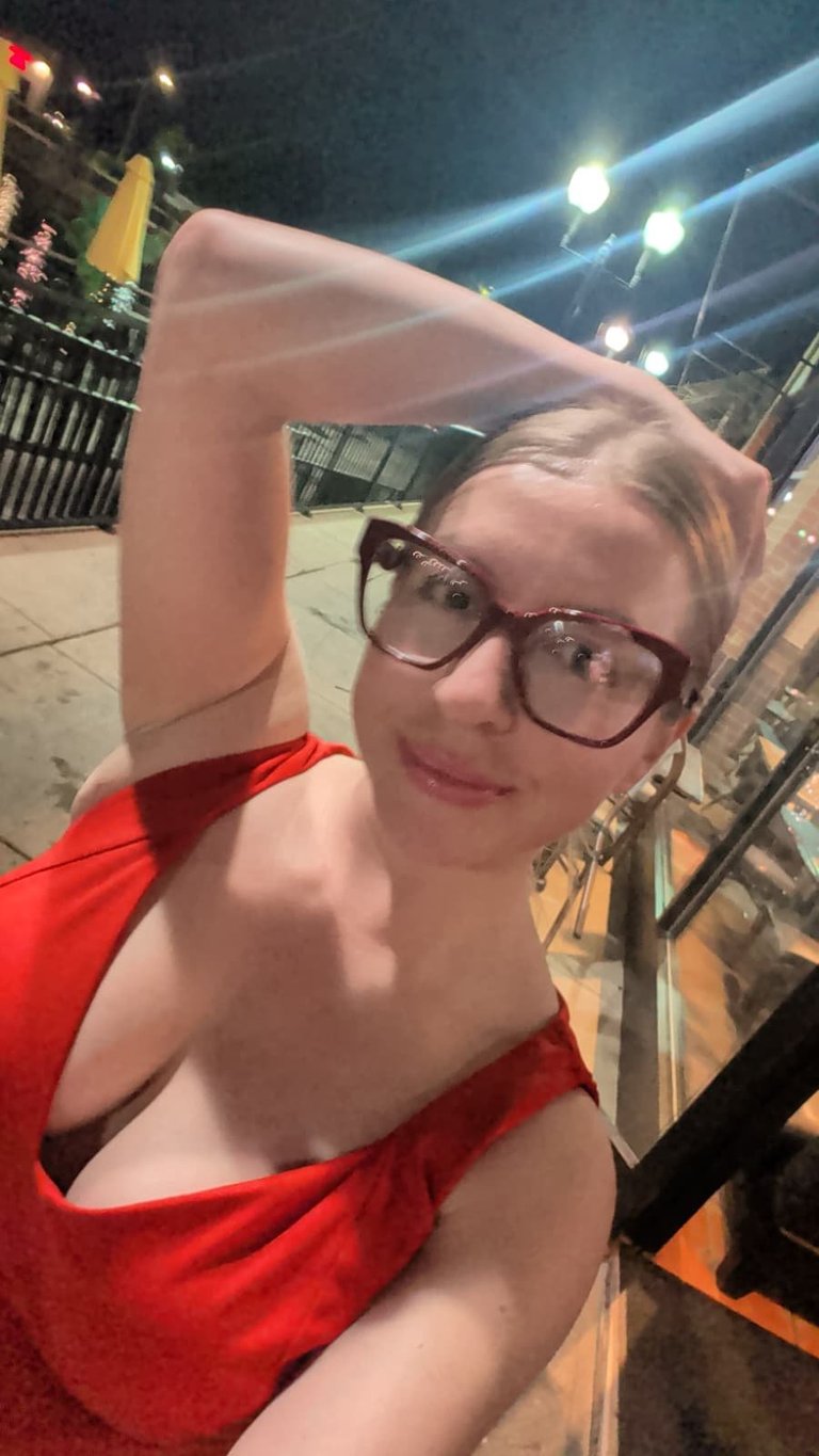 Person wearing glasses and red sleeveless top posing for selfie outdoors at night with lit street and city lights in background